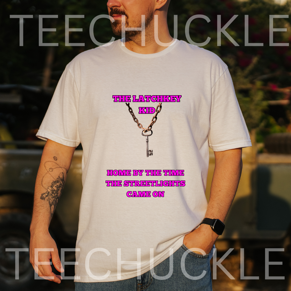 A man wearing a white graphic t-shirt with a pink print that reads 'THE LATCHKEY KID HOME BY THE TIME THE STREETLIGHTS CAME ON' with an illustration of a key and chain. The background is a blurred outdoor setting with warm sunset lighting.