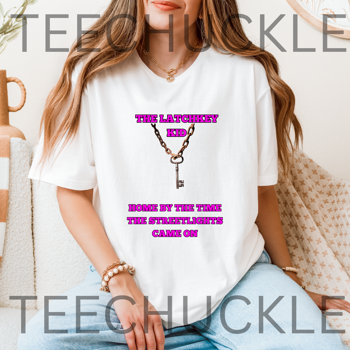 A woman wearing a white graphic t-shirt with a pink that reads 'THE LATCHKEY KID HOME BY THE TIME THE STREETLIGHTS CAME ON' around an illustration of a key.
