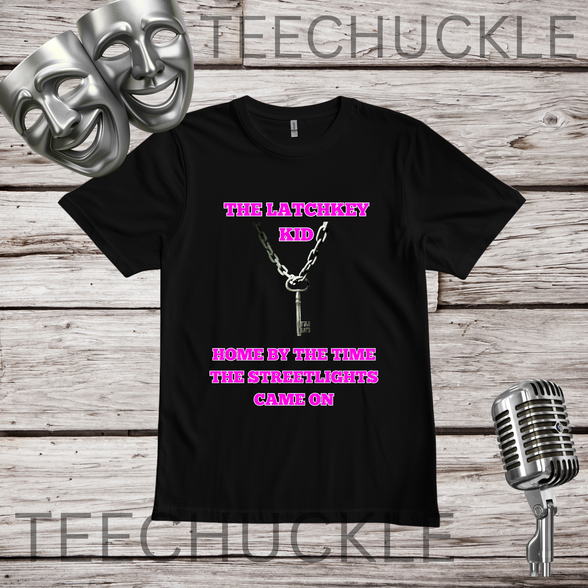 A black short-sleeve graphic T-shirt laid flat on a rustic wood background, with two silver theater masks, a vintage microphone. The shirt has pink text reading 'THE LATCHKEY KID' and 'HOME BY THE TIME THE STREETLIGHTS GAVE ON' with a silver keychain graphic in the center.