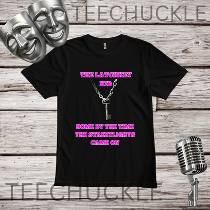 A black short-sleeve graphic T-shirt laid flat on a rustic wood background, with two silver theater masks, a vintage microphone. The shirt has pink text reading 'THE LATCHKEY KID' and 'HOME BY THE TIME THE STREETLIGHTS GAVE ON' with a silver keychain graphic in the center.