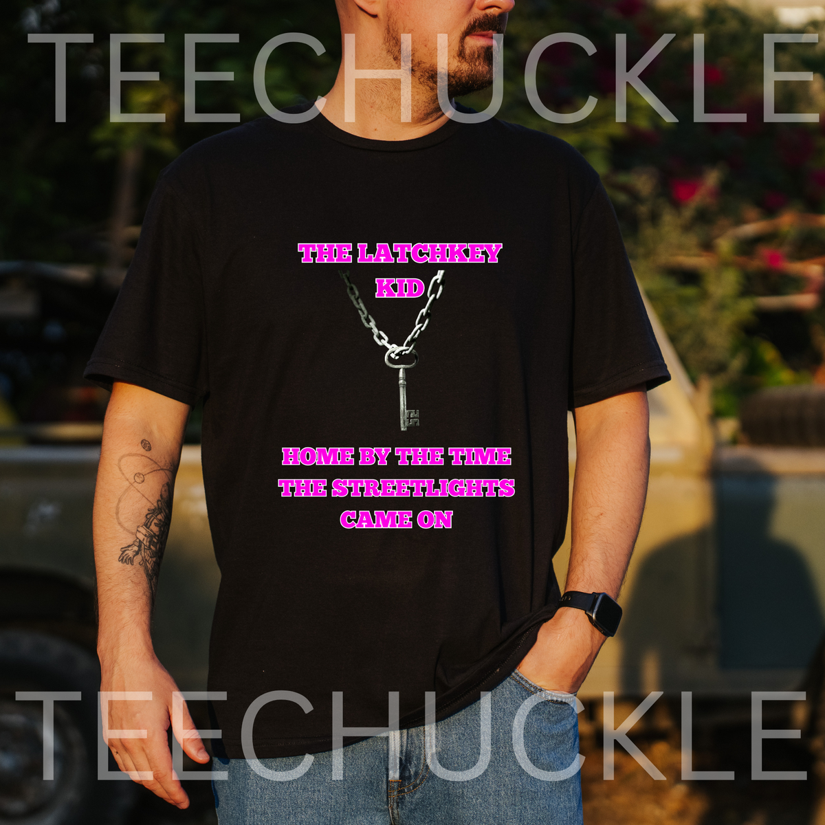 A man wearing a black graphic t-shirt with a pink and white design that reads 'TIME LATCHKEY KID' and 'HOME BY THE TIME THE STREETLIGHTS GAME ON'. The background is a blurred outdoor setting with a vehicle