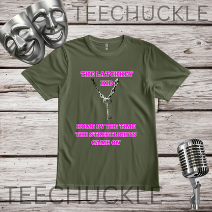 A green graphic t-shirt displayed on a rustic wood background, featuring a key and chain with the pink text 'THE LATCHKEY KID HOME BY THE TIME THE STREETLIGHTS CAME ON'. The image includes decorative props like silver theater masks and a vintage microphone