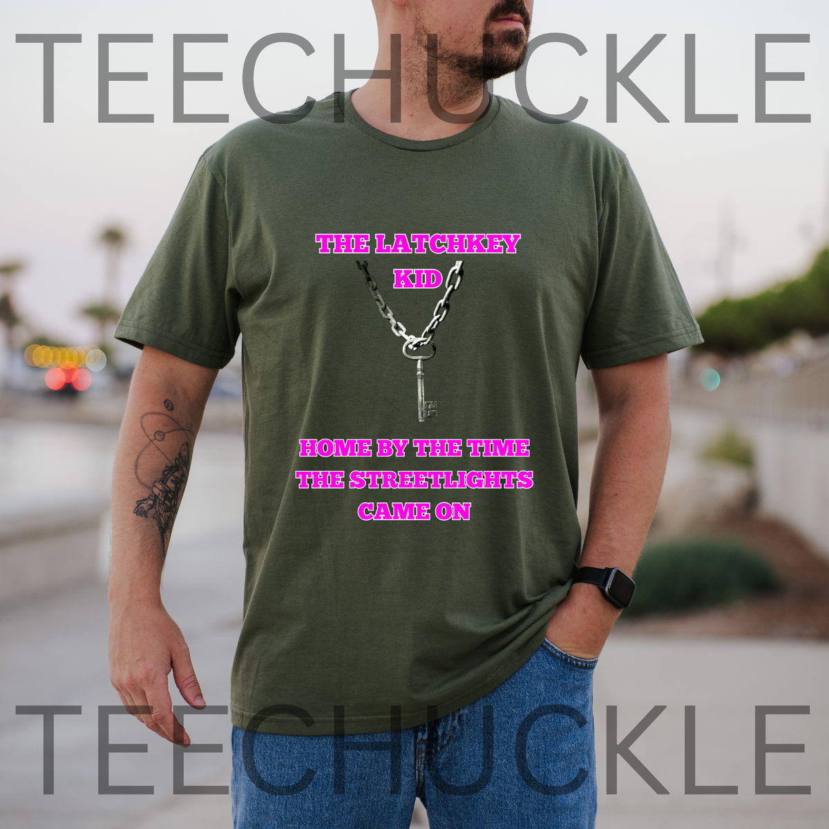 A man wearing a green graphic t-shirt with pink text reading 'THE LATCHKEY KID HOME BY THE TIME THE STREETLIGHTS GAVE ON' and a silver keychain graphic, set against a blurred outdoor background. Large 