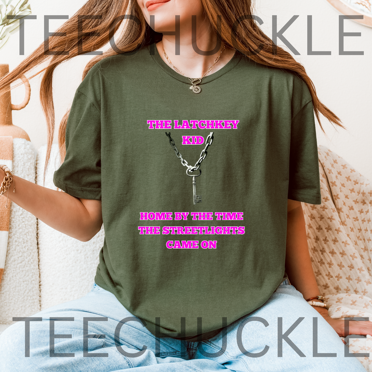 A woman wearing a green graphic t-shirt with a silver key illustration, sitting in a cozy indoor setting. The t-shirt features the text 'THE LATCHKEY KIDS' and 'HOME BY THE TIME THE STRENGTHS CAME ON'.