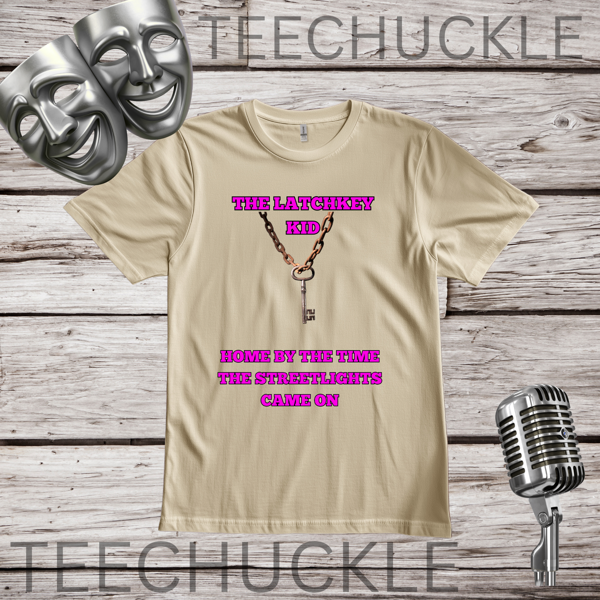 A sand graphic t-shirt displayed on a rustic wood background, featuring a key necklace design and pink text that reads 'THE LATCHKEY KID' and 'HOME BY THE TIME THE STREETLIGHTS CAME ON'. The shirt is styled with silver theater masks, a vintage microphone.