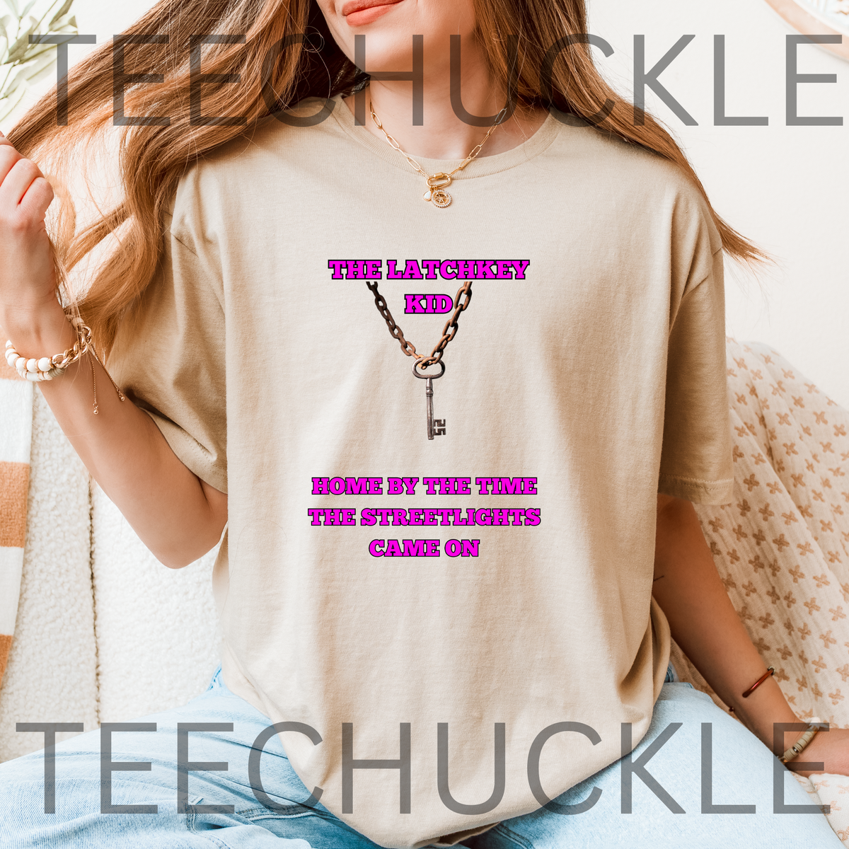 A woman wearing a sand graphic t-shirt with a key and chain design, sitting in a cozy indoor setting. The t-shirt features the text 'THE LATCHKEY KID' and 'HOME BY THE TIME THE STREETLIGHTS CAME ON' in pink around a key graphic.