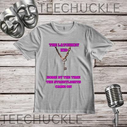 A gray graphic t-shirt featuring a keychain design with the text 'THE LATCHKEY KID' and 'HOME BY THE TIME THE STREETLIGHTS CAME ON', displayed on a rustic wood background with vintage props including a microphone, comedy mask.