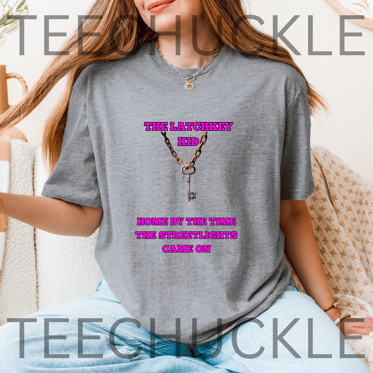 A woman wearing a gray graphic t-shirt with a pink text  reading 'THE LATCHKEY KID HOME BY THE TIME THE STREETLIGHTS CAME ON' around a key graphic. 