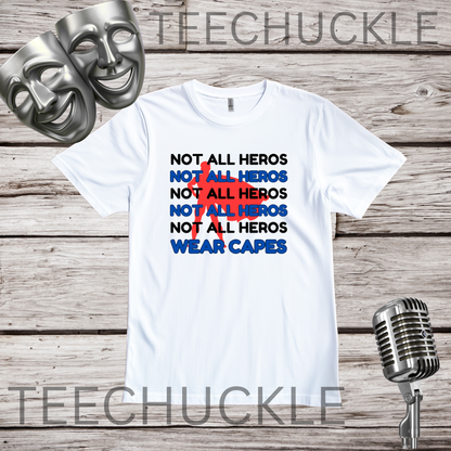 A white graphic t-shirt displayed on a rustic wood background, flanked by two silver theatrical masks on the left and a vintage silver microphone on the right. The t-shirt features a text graphic reading 'NOT ALL HEROS' repeated in black, blue, and red, with 'WEAR CAPES' in blue.