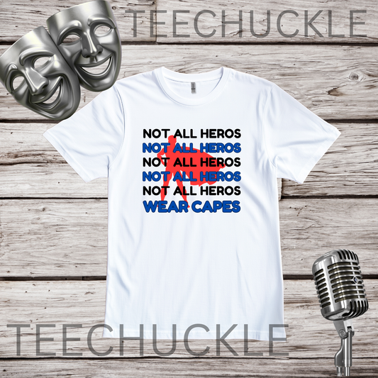 A white graphic t-shirt displayed on a rustic wood background, flanked by two silver theatrical masks on the left and a vintage silver microphone on the right. The t-shirt features a text graphic reading 'NOT ALL HEROS' repeated in black, blue, and red, with 'WEAR CAPES' in blue.