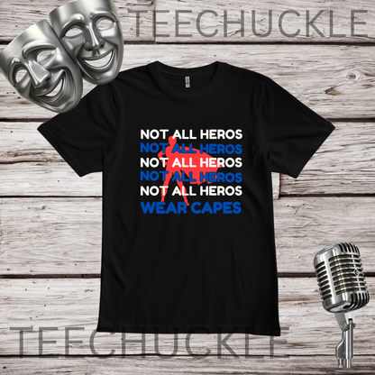 A black short-sleeve graphic T-shirt laid flat on a rustic wood background, with theatrical masks and a vintage microphone as props. The shirt has repeated white and blue text reading 'NOT ALL HEROS' and blue text reading 'WEAR CAPES' in the center.