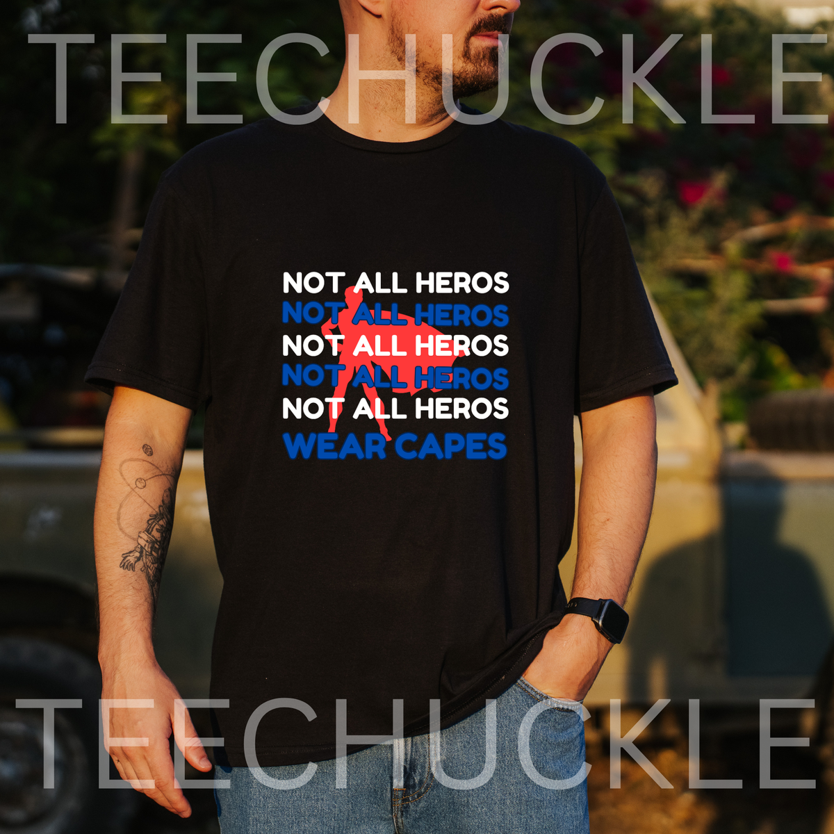 A man wearing a black graphic t-shirt with the slogan 'NOT ALL HEROS' repeated in white and blue, with a red superhero behind it, and finished with 'WEAR CAPES' in blue. 
