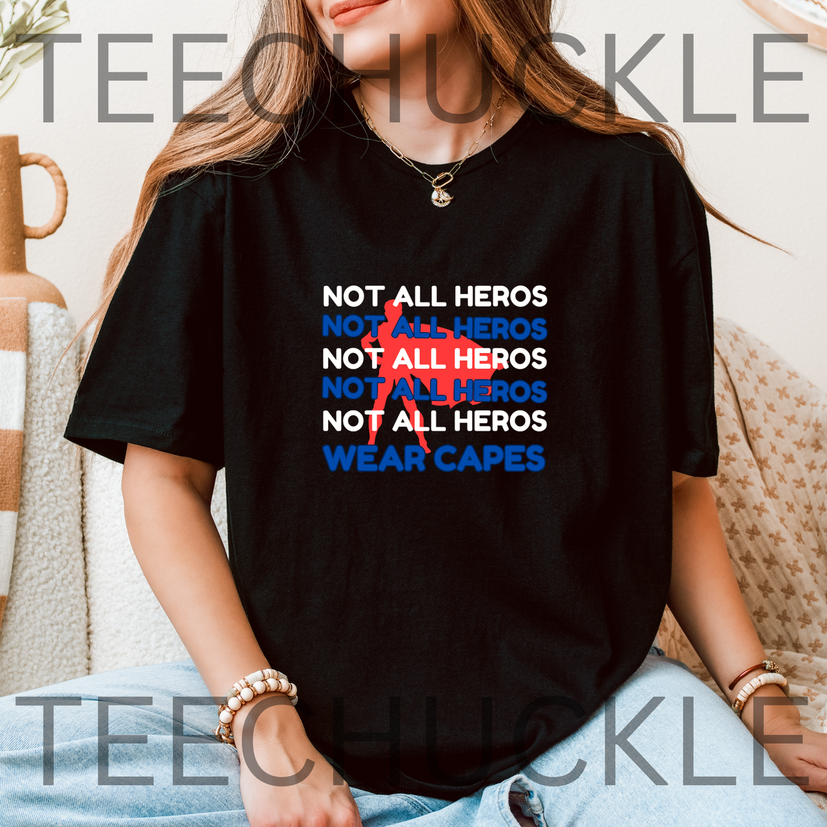 A woman wearing a black graphic T-shirt with the text 'NOT ALL HEROS' repeated in white and blue, with a red superhero behind, and "wear capes" at the bottom, sitting indoors.