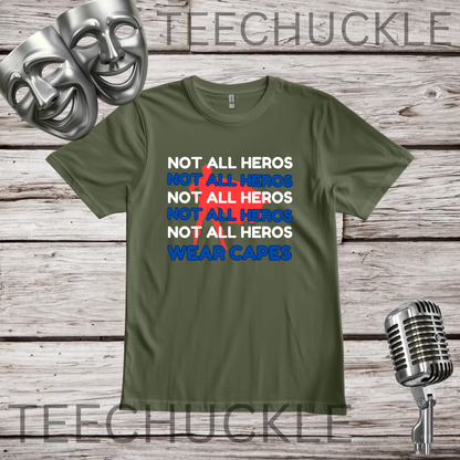 A green graphic t-shirt displayed on a rustic wooden background, featuring a text design that reads 'NOT ALL HEROS' in white and blue, with a red superhero behind it, and finished with 'WEAR CAPES' in blue. The t-shirt is framed by silver comedy masks on the top left, a vintage microphone on the bottom right.