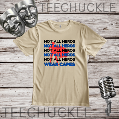 A sand graphic t-shirt displayed on a rustic wood background, flanked by two silver comedy masks on the top left and a vintage silver microphone on the bottom right. The t-shirt features a stacked text graphic reading 'NOT ALL HEROS' in black, and blue, with a red superhero behind it
