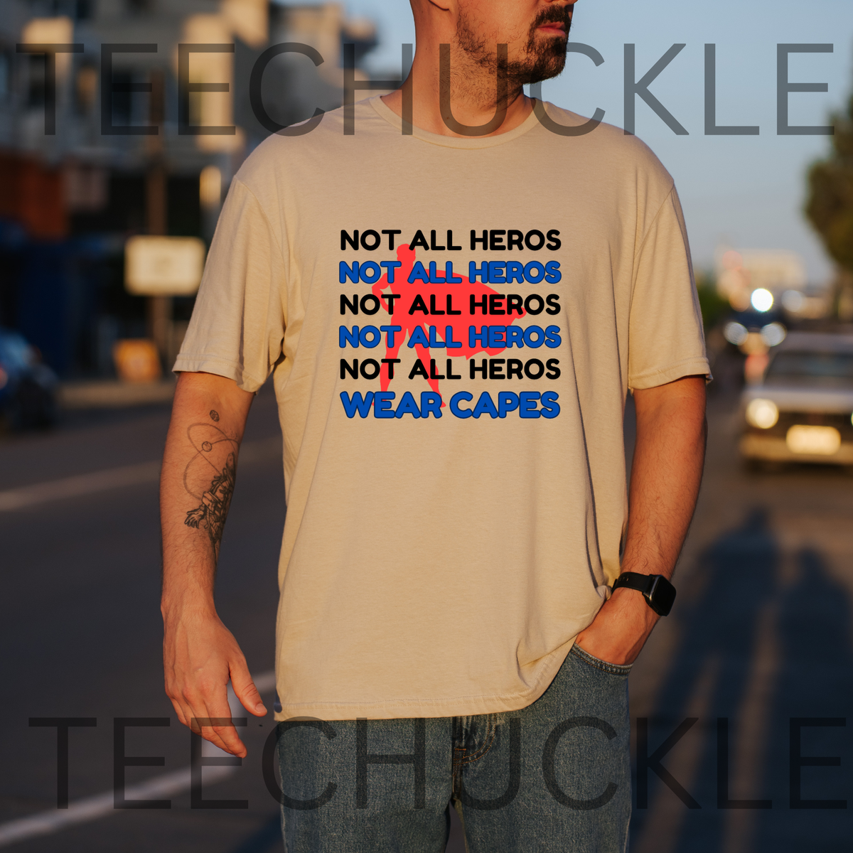 A man wearing a sand graphic t-shirt with the text 'NOT ALL HEROS NOT ALL HEROS NOT ALL HEROS NOT ALL HEROS WEAR CAPES' in black and blue, with a red superhero behind it, standing on a street at sunset. 