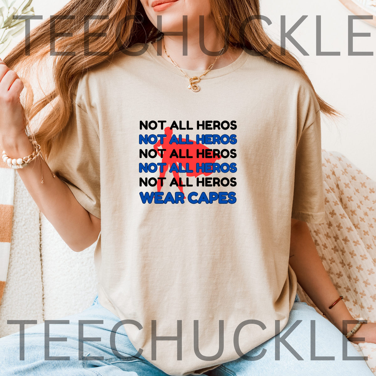 A woman wearing a sand graphic T-shirt with the text 'NOT ALL HEROS' repeated in black and blue, with a red superhero behind it, and "wear capes" at the bottom