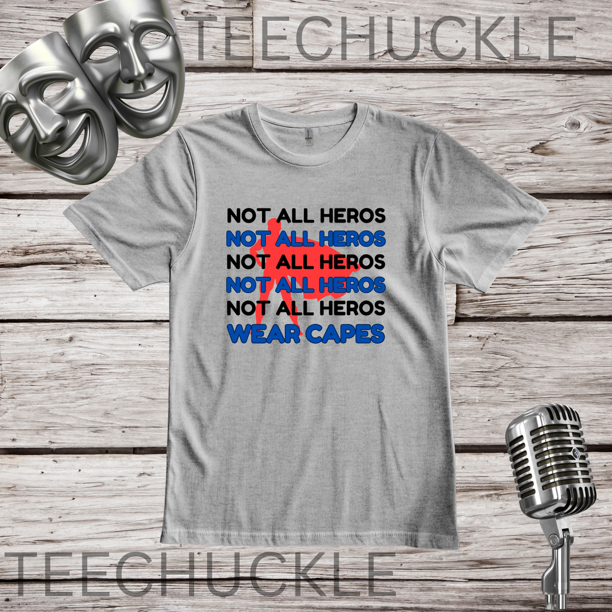 A gray graphic t-shirt displayed on a rustic wood background, featuring a 'NOT ALL HEROS' in black and blue text with a red superhero behind it and the text "wear capes" at the bottom. The shirt is styled with theatrical masks, a vintage microphone.