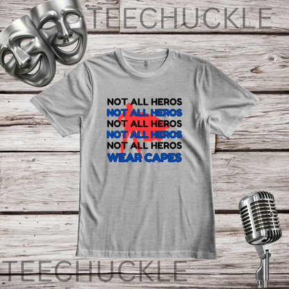 A gray graphic t-shirt displayed on a rustic wood background, featuring a 'NOT ALL HEROS' in black and blue text with a red superhero behind it and the text "wear capes" at the bottom. The shirt is styled with theatrical masks, a vintage microphone.