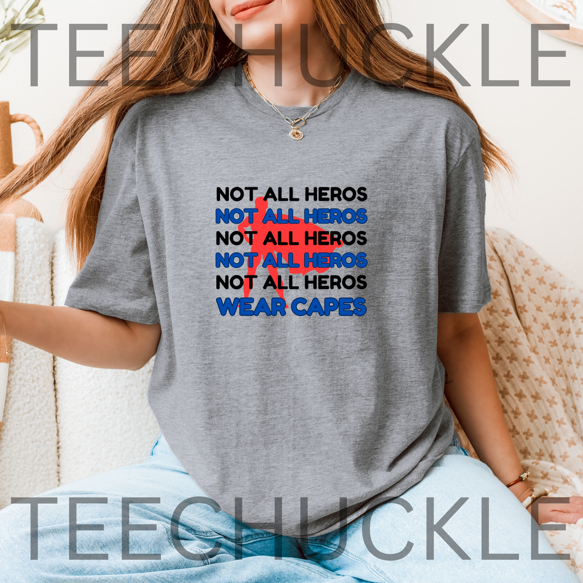 A woman wearing a gray graphic t-shirt with the text 'NOT ALL HEROS' repeated in black and blue, with a red superhero behind it and "waer capes" at the bottom