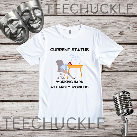 A white graphic t-shirt displayed flat on a rustic wood background, featuring a cartoon illustration of a person sleeping at a desk with the text 'CURRENT STATUS WORKING HARD AT HARDLY WORKING' with decorative silver comedy masks on the left and a vintage microphone on the right.
