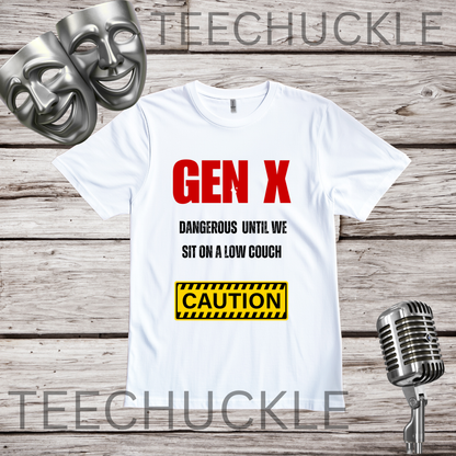A white t-shirt displayed on a rustic wood background, featuring a red and black text design that reads 'GEN X DANGEROUS UNTIL WE SIT ON A LOW COUCH' with a yellow caution box below. Two silver comedy masks are on the top left, and a vintage silver microphone is on the bottom right.