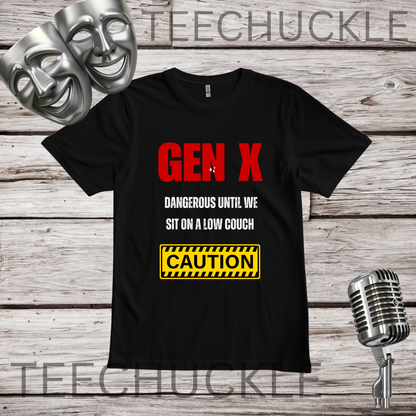 A black short-sleeve graphic T-shirt laid flat on a rustic wood plank background, with two silver theater masks and a vintage microphone as props. The shirt has large red 'GEN X' text, smaller white text reading 'DANGEROUS UNTIL WE SIT ON A LOW COUCH,' and a yellow caution-style box with black stripes and the word 'CAUTION.