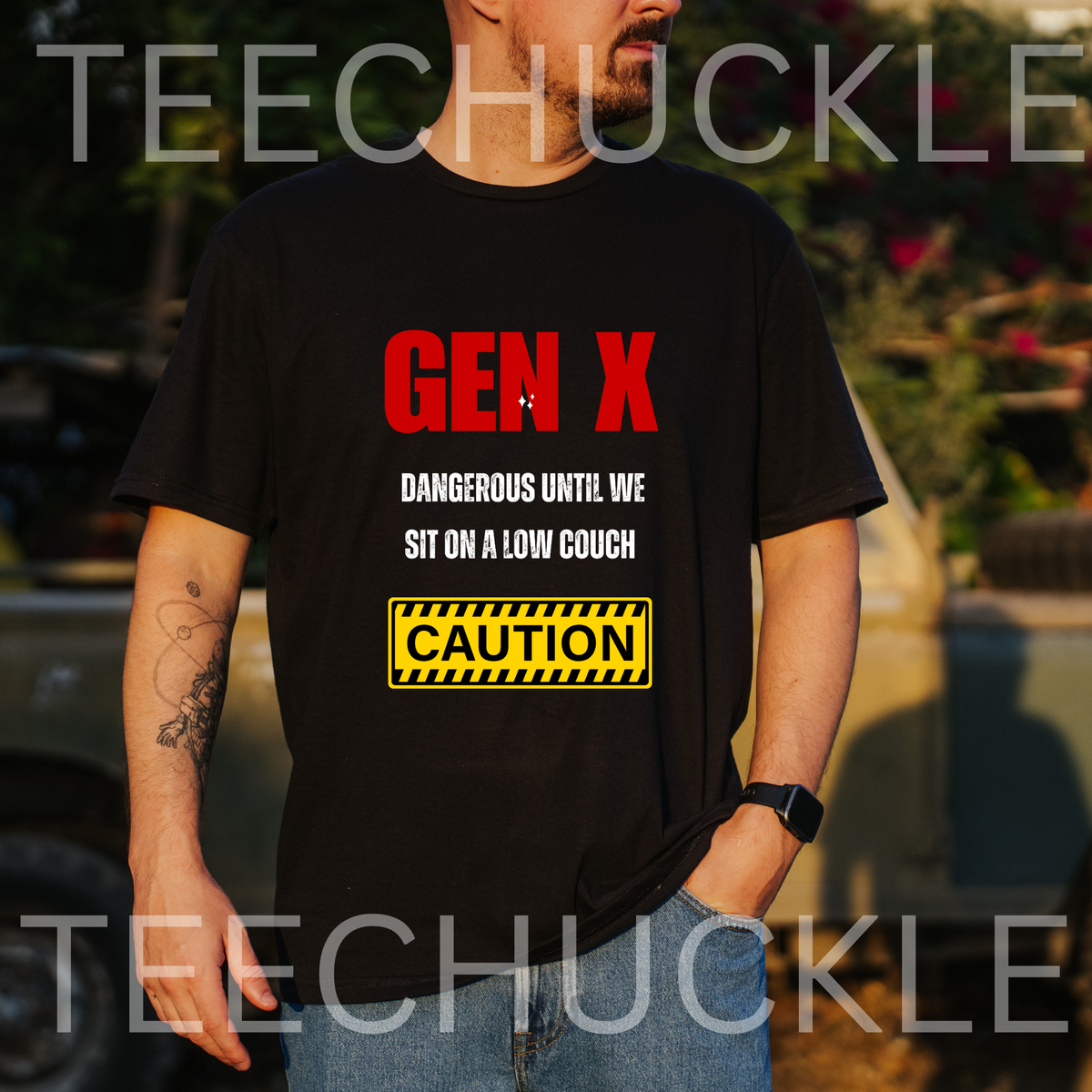 A man wearing a black graphic t-shirt with the text 'GEN X DANGEROUS UNTIL WE SIT ON A LOW COUCH' and a yellow caution sign graphic. The background is an outdoor setting with a blurred vehicle and foliage.