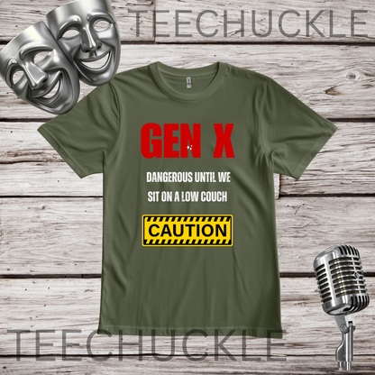 A green graphic t-shirt displayed on a rustic wood background with silver theater masks and a vintage microphone. The t-shirt features the text 'GEN X' in red, 'DANGEROUS UNTIL WE SIT ON A LOW COUCH' in white, and a yellow 'CAUTION' warning label graphic