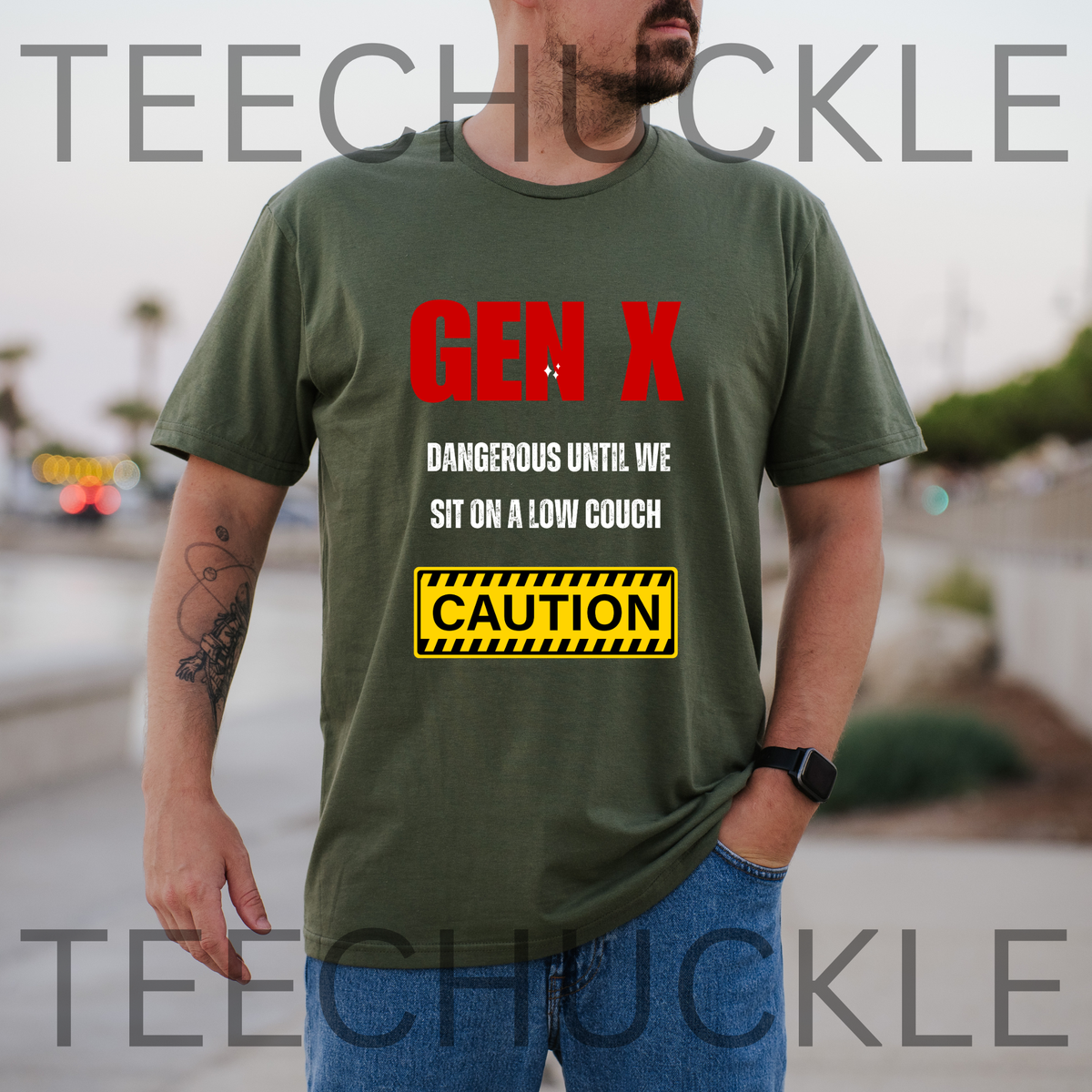 A man wearing a green graphic t-shirt with the text 'GEN X DANGEROUS UNTIL WE SIT ON A LOW COUCH' and a yellow 'CAUTION' sign graphic. The background is a blurred outdoor street scene.