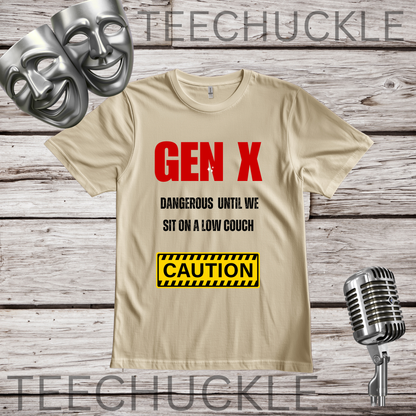 A sand graphic t-shirt displayed on a rustic wood background, featuring the text 'GEN X DANGEROUS UNTIL WE SIT ON A LOW COUCH' in red and black, and a yellow 'CAUTION' box graphic, and styled with silver comedy masks and a vintage microphone.