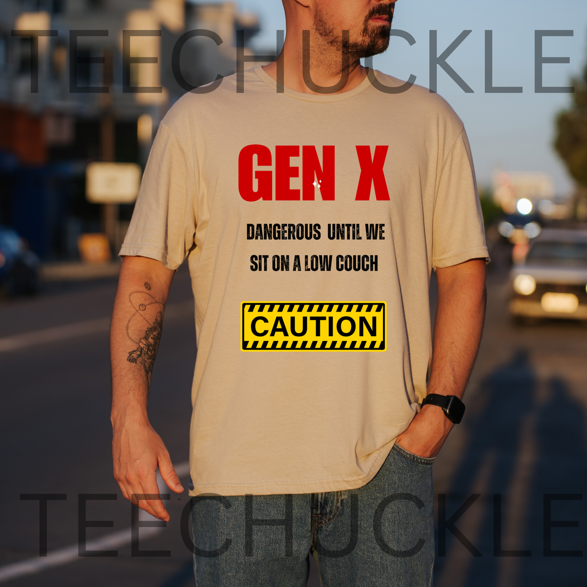 A man wearing a sand graphic t-shirt with a 'Gen X' warning label design, standing outdoors on a street at sunset. The t-shirt features red and black text reading 'GEN X DANGEROUS UNTIL WE SIT ON A LOW COUCH' and a yellow 'CAUTION' box.