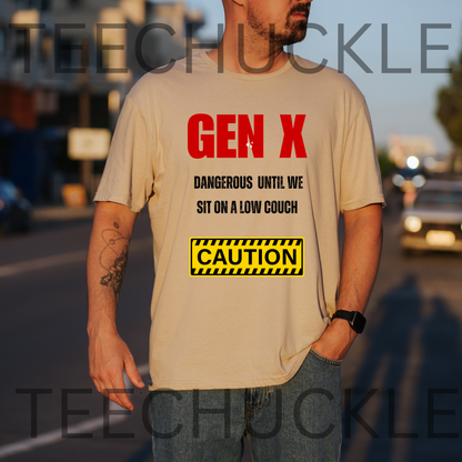 A man wearing a sand graphic t-shirt with a 'Gen X' warning label design, standing outdoors on a street at sunset. The t-shirt features red and black text reading 'GEN X DANGEROUS UNTIL WE SIT ON A LOW COUCH' and a yellow 'CAUTION' box.