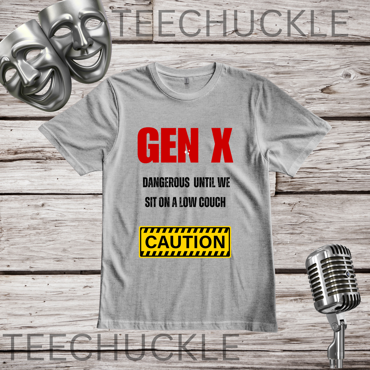 A gray graphic t-shirt displayed flat on a rustic wooden background, featuring the text 'GEN X DANGEROUS UNTIL WE SIT ON A LOW GOUCH' in red and black, and a yellow 'CAUTION' box graphic. Silver comedy masks and a vintage microphone are placed in the corners.