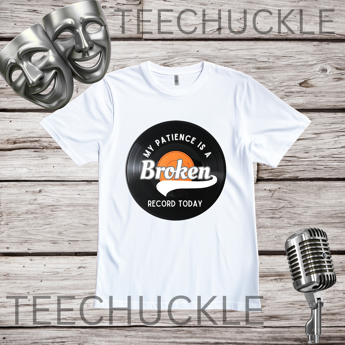 A white short-sleeve graphic T-shirt laid flat on a rustic wood plank background, with two silver comedy masks in the upper left and a vintage-style microphone in the lower right. The shirt has a vinyl record graphic reading 'My Patience Is a Broken Record Today.