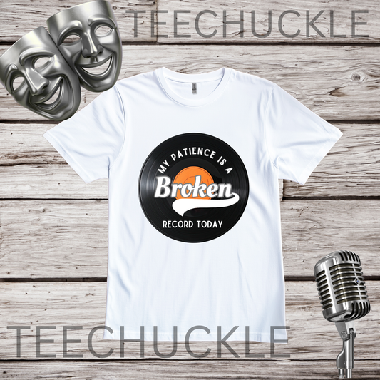 A white short-sleeve graphic T-shirt laid flat on a rustic wood plank background, with two silver comedy masks in the upper left and a vintage-style microphone in the lower right. The shirt has a vinyl record graphic reading 'My Patience Is a Broken Record Today.