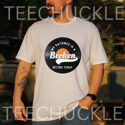 A man wearing a white graphic t-shirt with a vinyl record circular that reads 'MY PATIENCE IS A BROKEN RECORD TODAY'. The background is a blurred outdoor setting with a vehicle