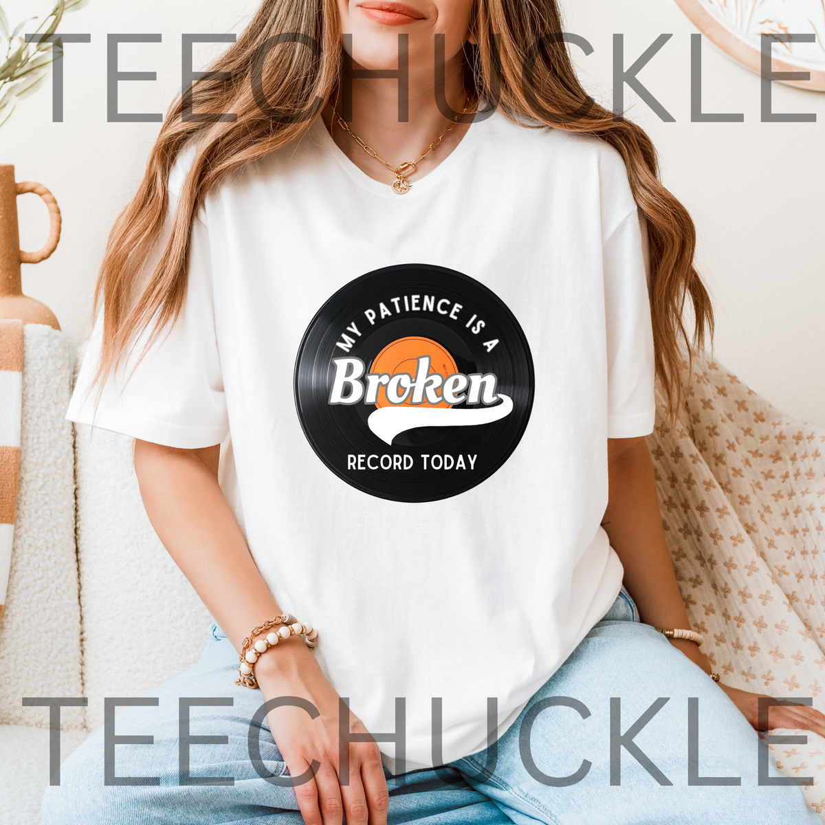 A woman wearing a white graphic t-shirt with a vinyl record that reads 'MY PATIENCE IS A BROKEN RECORD TODAY' and is set in a cozy indoor environment.