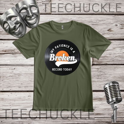 An green crewneck t-shirt with a vinyl record graphic on the chest that reads 'MY PATIENCE IS A BROKEN RECORD TODAY' in white. The shirt is displayed on a rustic wooden background with silver theater masks and a vintage microphone prop.