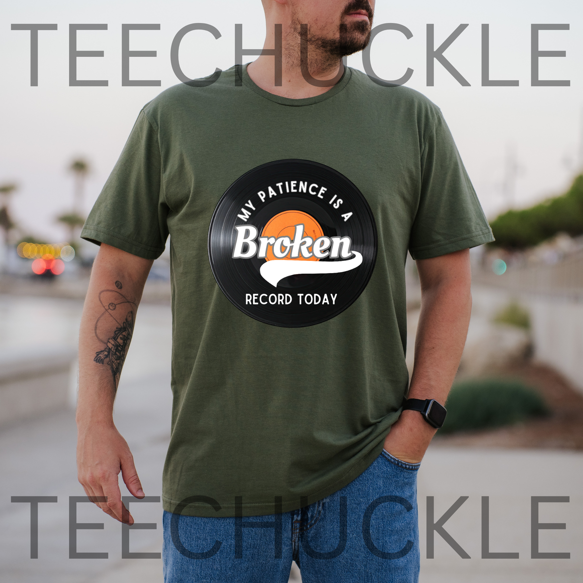A man wearing an green graphic t-shirt with a circular vinyl record design that reads 'MY PATIENCE IS A Broken RECORD TODAY'. The background is a blurred outdoor street scene.