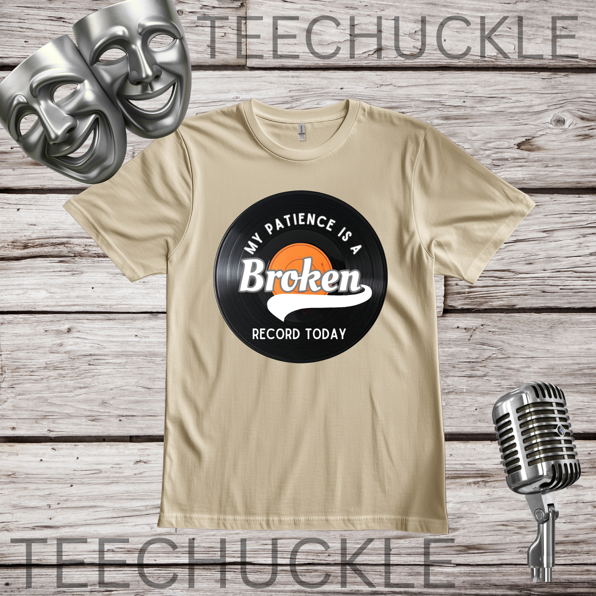 A SAND graphic t-shirt displayed on a rustic wood background, featuring a vinyl record graphic that reads 'MY PATIENCE IS A BROKEN RECORD TODAY'. The image includes decorative props like silver comedy masks, a vintage microphone.