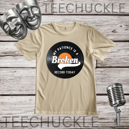 A SAND graphic t-shirt displayed on a rustic wood background, featuring a vinyl record graphic that reads 'MY PATIENCE IS A BROKEN RECORD TODAY'. The image includes decorative props like silver comedy masks, a vintage microphone.