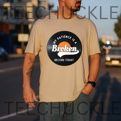 A man wearing a sand t-shirt with a vinyl record graphic that reads 'MY PATIENCE IS A BROKEN RECORD TODAY'. The background is a blurred street scene at sunset