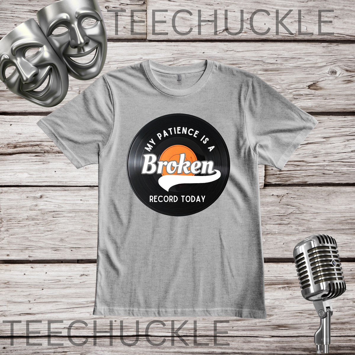 A gray t-shirt laid flat on a rustic wood background, featuring a vinyl record graphic and the words 'MY PATIENCE IS A BROKEN RECORD TODAY'. The shirt is styled with comedy masks in the top-left corner and a vintage microphone in the bottom-right corner.