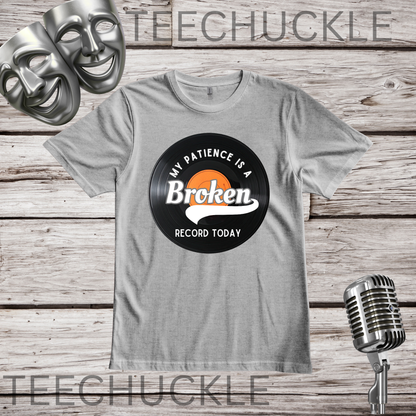 A gray t-shirt laid flat on a rustic wood background, featuring a vinyl record graphic and the words 'MY PATIENCE IS A BROKEN RECORD TODAY'. The shirt is styled with comedy masks in the top-left corner and a vintage microphone in the bottom-right corner.