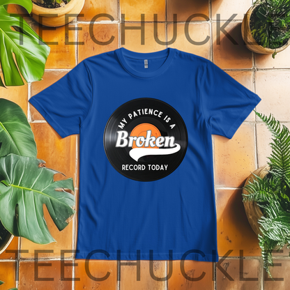 A royal blue short-sleeve t-shirt laid flat on a terracotta tile surface, surrounded by green plants. The shirt features a vinyl record graphic and white text that reads 'MY PATIENCE IS A BROKEN RECORD TODAY'.