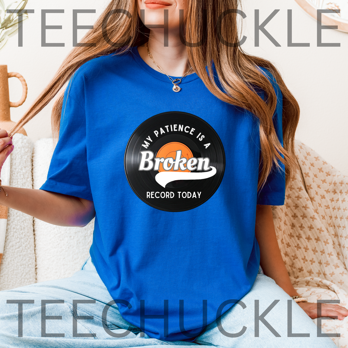A woman wearing a royal blue t-shirt with a vinyl record graphic with text that reads 'MY PATIENCE IS A Broken RECORD TODAY', and the model is sitting on a beige patterned couch.