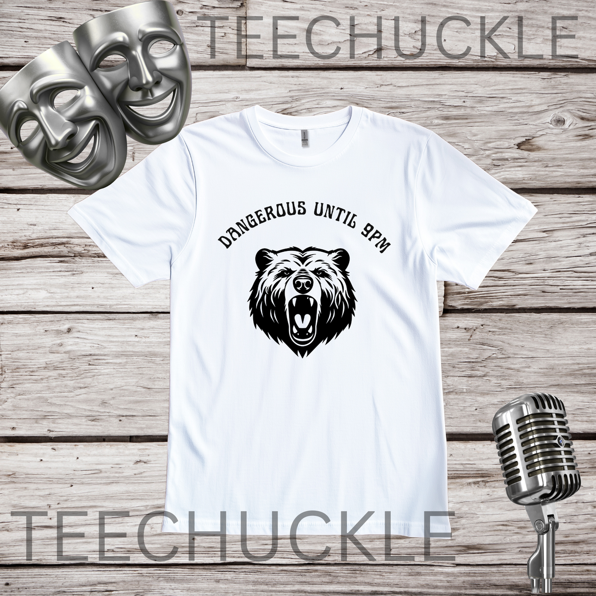A white short-sleeve graphic T-shirt laid flat on a rustic wood plank background, featuring a black bear head illustration and the text 'DANGEROUS UNTIL 9PM' above it. Two silver theatrical masks and a vintage-style microphone are placed around the shirt as props.