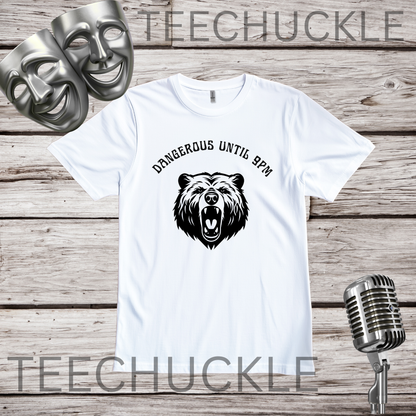 A white short-sleeve graphic T-shirt laid flat on a rustic wood plank background, featuring a black bear head illustration and the text 'DANGEROUS UNTIL 9PM' above it. Two silver theatrical masks and a vintage-style microphone are placed around the shirt as props.
