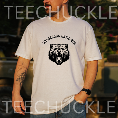 A man wearing a white crewneck t-shirt with a black bear graphic and the text 'DANGEROUS UNTIL 9PM' on the front, set against a blurred outdoor background. 